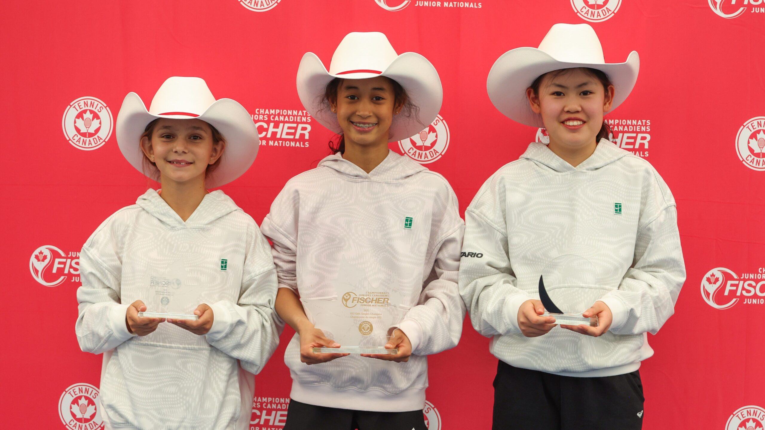 Three girls from the U12 junior nationals, wearing stetsons, hold up their trophies.
